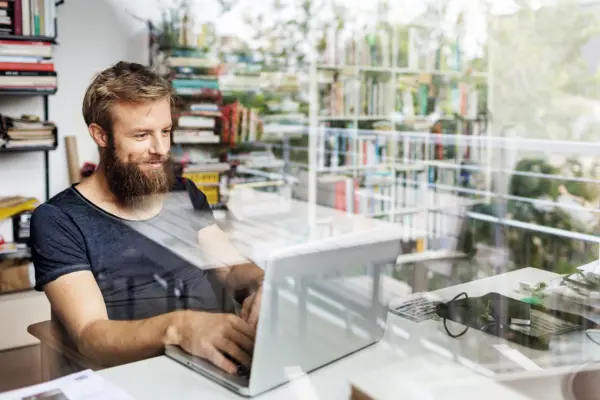 Ein junger Mann mit Vollbart arbeitet konzentriert an einem Laptop. Das Foto wurde durch eine Glasscheibe aufgenommen, in der sich Bücherregale und die Umgebung spiegeln.