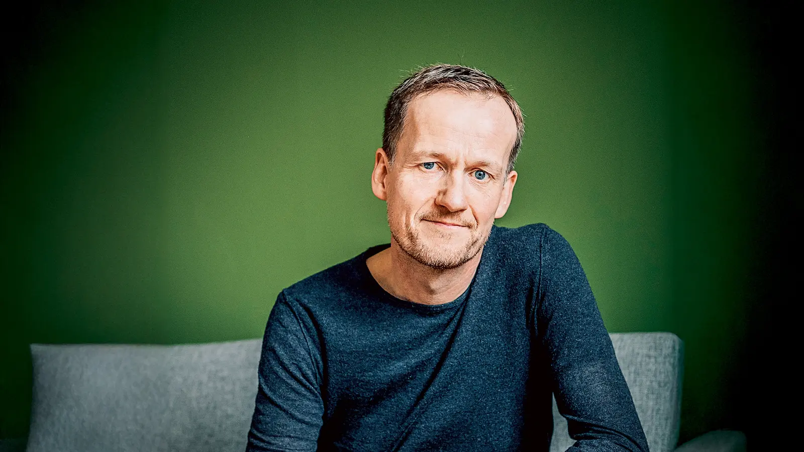 Ein Mann mittleren Alters mit kurzen, hellbraunen Haaren und hellem Teint sitzt auf einem grauen Sofa vor einer grünen Wand. Er trägt ein dunkelblaues Langarmshirt und eine blaue Jeans. Der Mann blickt mit einem leichten Lächeln in die Kamera und hat eine entspannte Körperhaltung, wobei er mit einer Hand auf seinem Knie ruht.