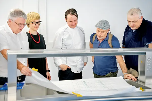 Fünf Personen begutachten gemeinsam Baupläne und besprechen ein laufendes Bauprojekt.