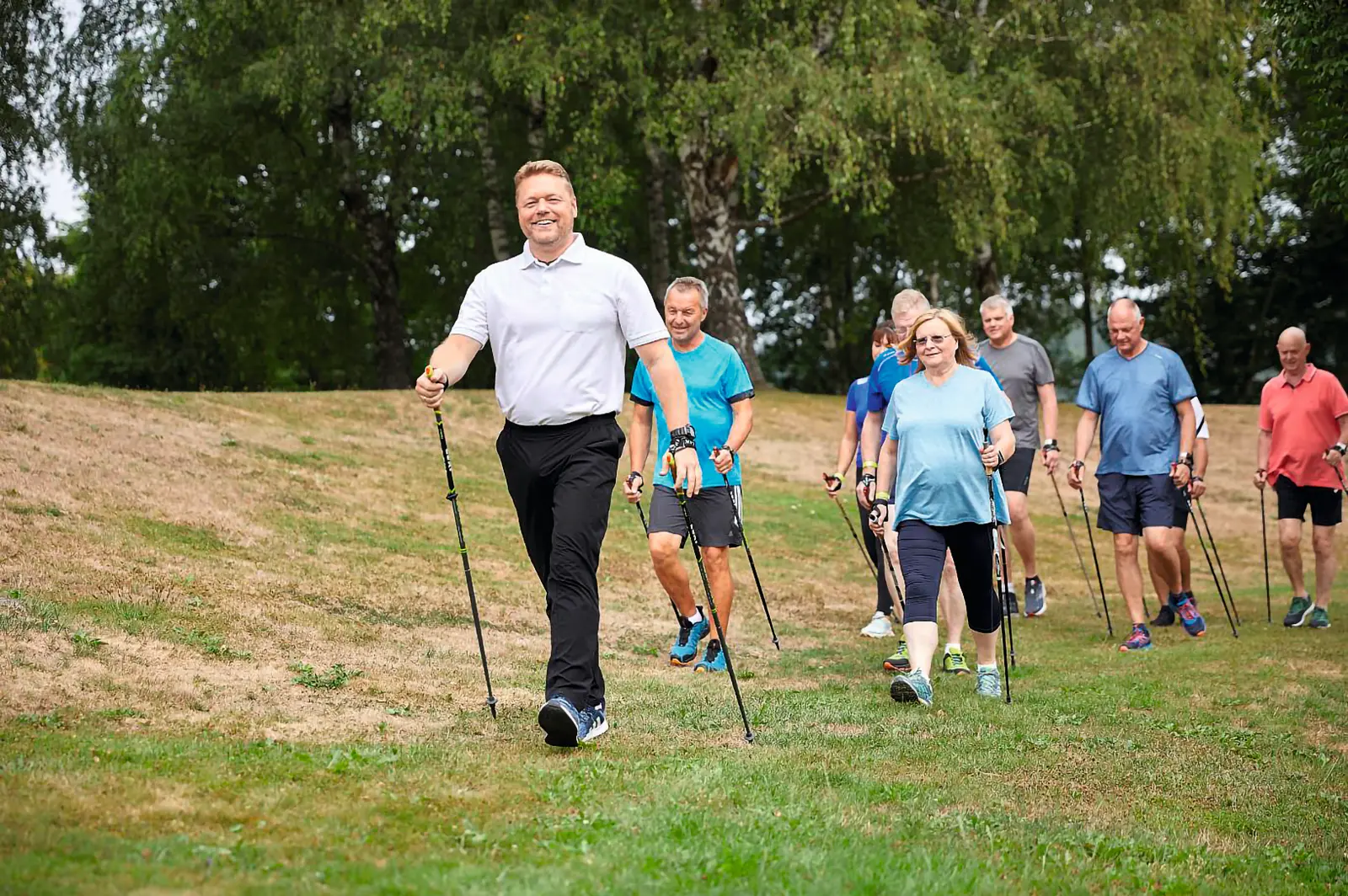 Eine Gruppe von Männern und Frauen praktiziert Nordic Walking auf einer grünen Wiese. Ein lächelnder Trainer in weißem Poloshirt führt die motivierte Gruppe an, alle halten Nordic-Walking-Stöcke in den Händen.