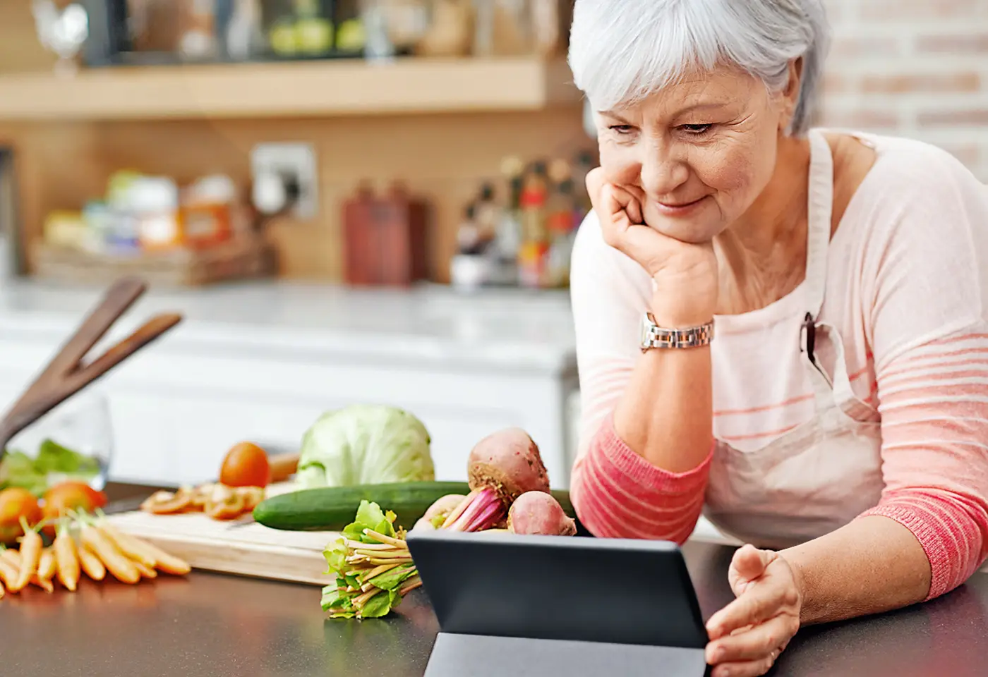 Eine ältere Frau steht in der Küche, schaut auf ein Tablet und bereitet frisches Gemüse zum Kochen vor.