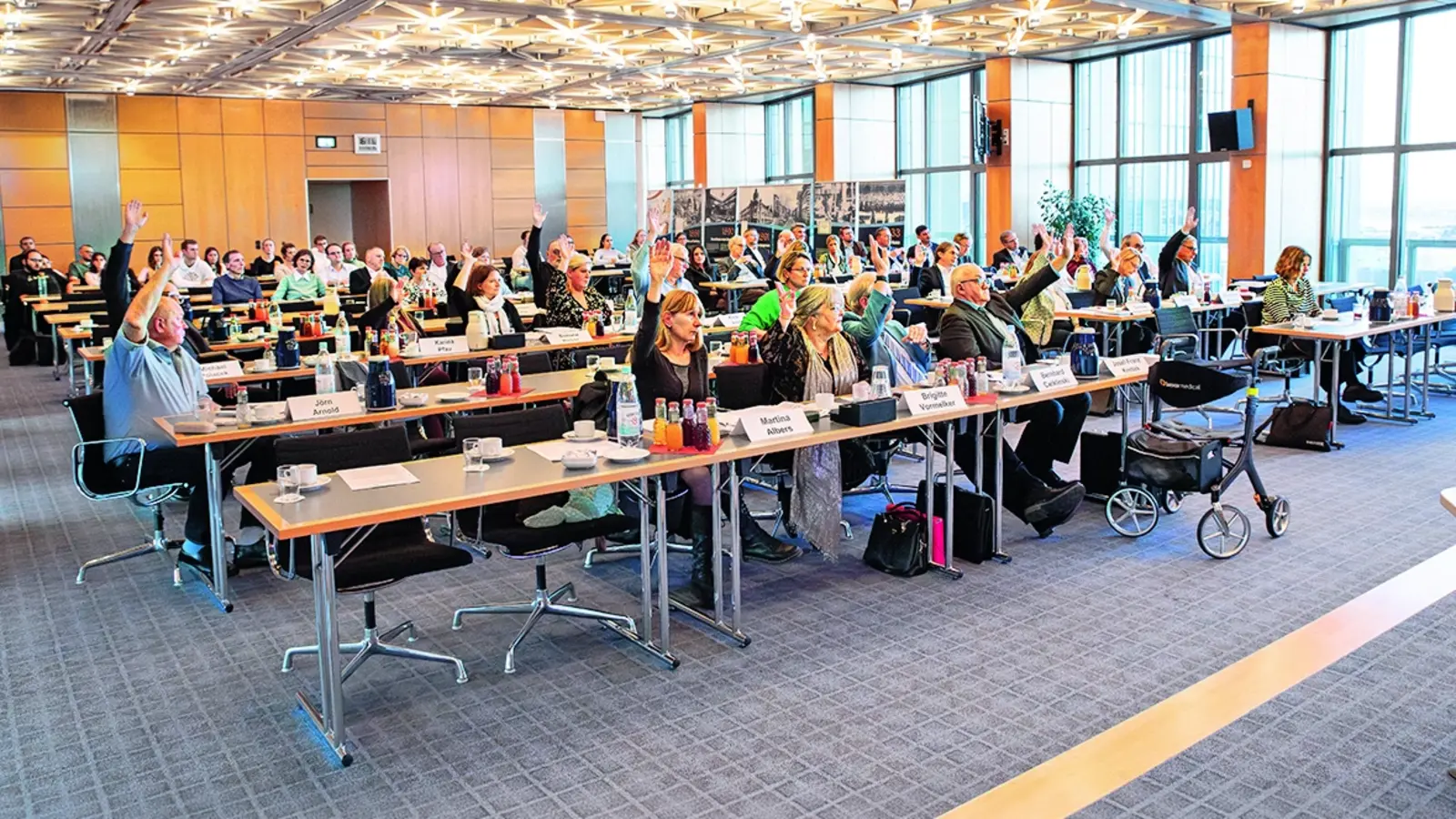 Mehrere Menschen befinden sich in einem Saal und sitzen am Tisch um für etwas abzustimmen. 