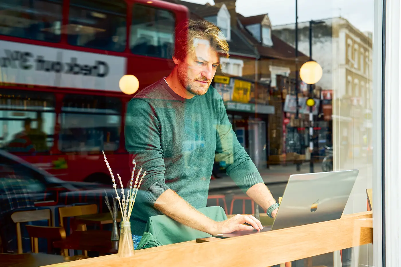 ChatGPT:  Ein Mann sitzt in einem Café und arbeitet an einem Laptop. Durch das Fenster ist eine Londoner Straße mit einem roten Doppeldeckerbus zu sehen. Die Szene wirkt urban und modern.