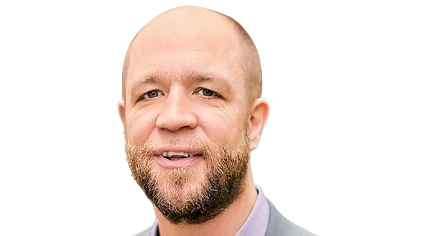 Porträt eines Mannes mit kurzem, glattem Haar und einem Vollbart. Er trägt ein graues Sakko und ein lila Hemd. Der Mann lächelt leicht und schaut freundlich in die Kamera.