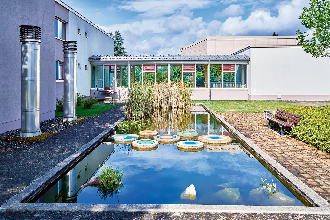 Innenhof einer modernen Rehaklinik mit rechteckigem Wasserbecken, in dem runde, bunt bemalte Trittsteine schwimmen. In der Mitte wachsen hohe Schilfpflanzen. Links steht ein flaches Gebäude mit großen Fenstern, rechts eine Holzbank neben einem gepflasterten Weg. Der Himmel ist blau mit einzelnen Wolken.