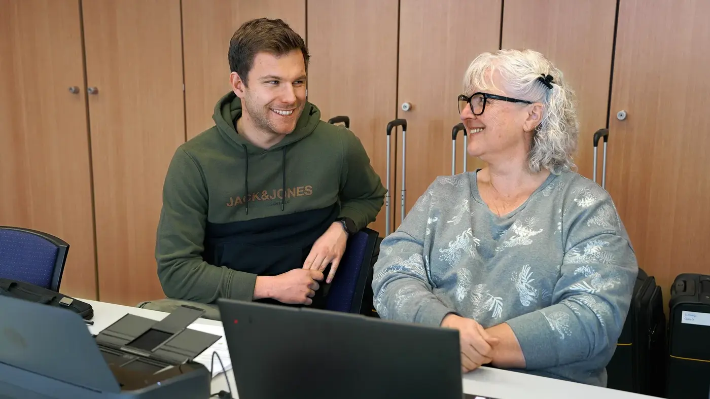 Ein junger Mann und eine ältere Frau sitzen am Tisch und schauen sich erfreut gegenseitig an.