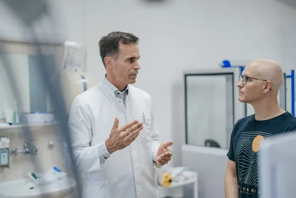 Das Bild zeigt zwei Männer in einem klinischen Umfeld. Einer trägt einen weißen Arztkittel und spricht gestikulierend, während der andere, mit einer Glatze und einer Brille, aufmerksam zuhört. Der Mann im Kittel steht links, während der andere rechts im Bild zu sehen ist. Die Szene ist in einer modernen und gut beleuchteten Umgebung aufgenommen.