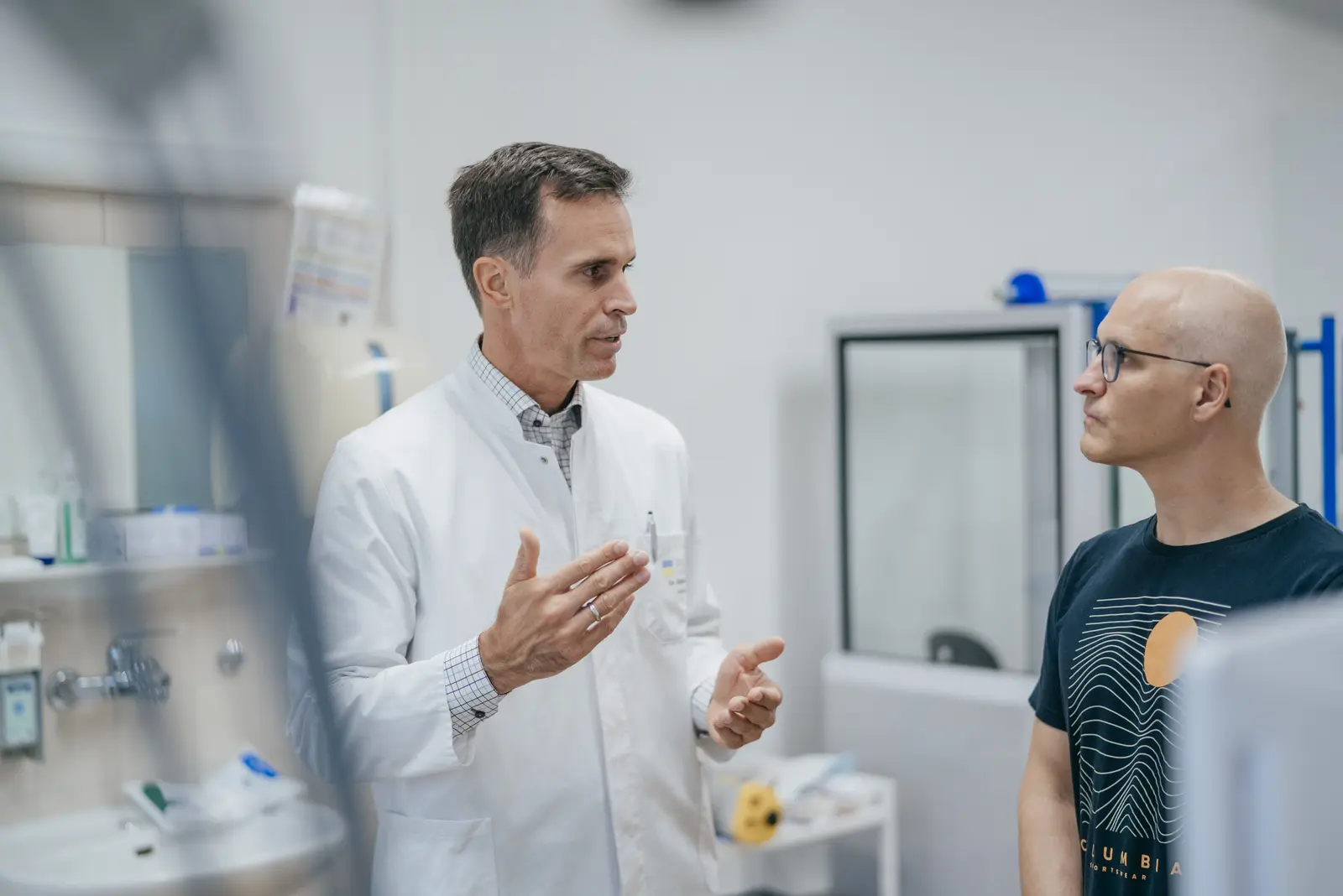 Das Bild zeigt zwei Männer in einem klinischen Umfeld. Einer trägt einen weißen Arztkittel und spricht gestikulierend, während der andere, mit einer Glatze und einer Brille, aufmerksam zuhört. Der Mann im Kittel steht links, während der andere rechts im Bild zu sehen ist. Die Szene ist in einer modernen und gut beleuchteten Umgebung aufgenommen.