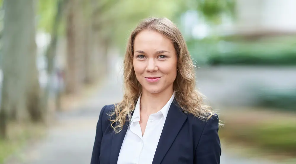 Porträtaufnahme einer Frau im Business-Outfit (dunkles Sakko, weiße Bluse), die freundlich in die Kamera blickt. Im Hintergrund ist eine verschwommene Allee erkennbar.
