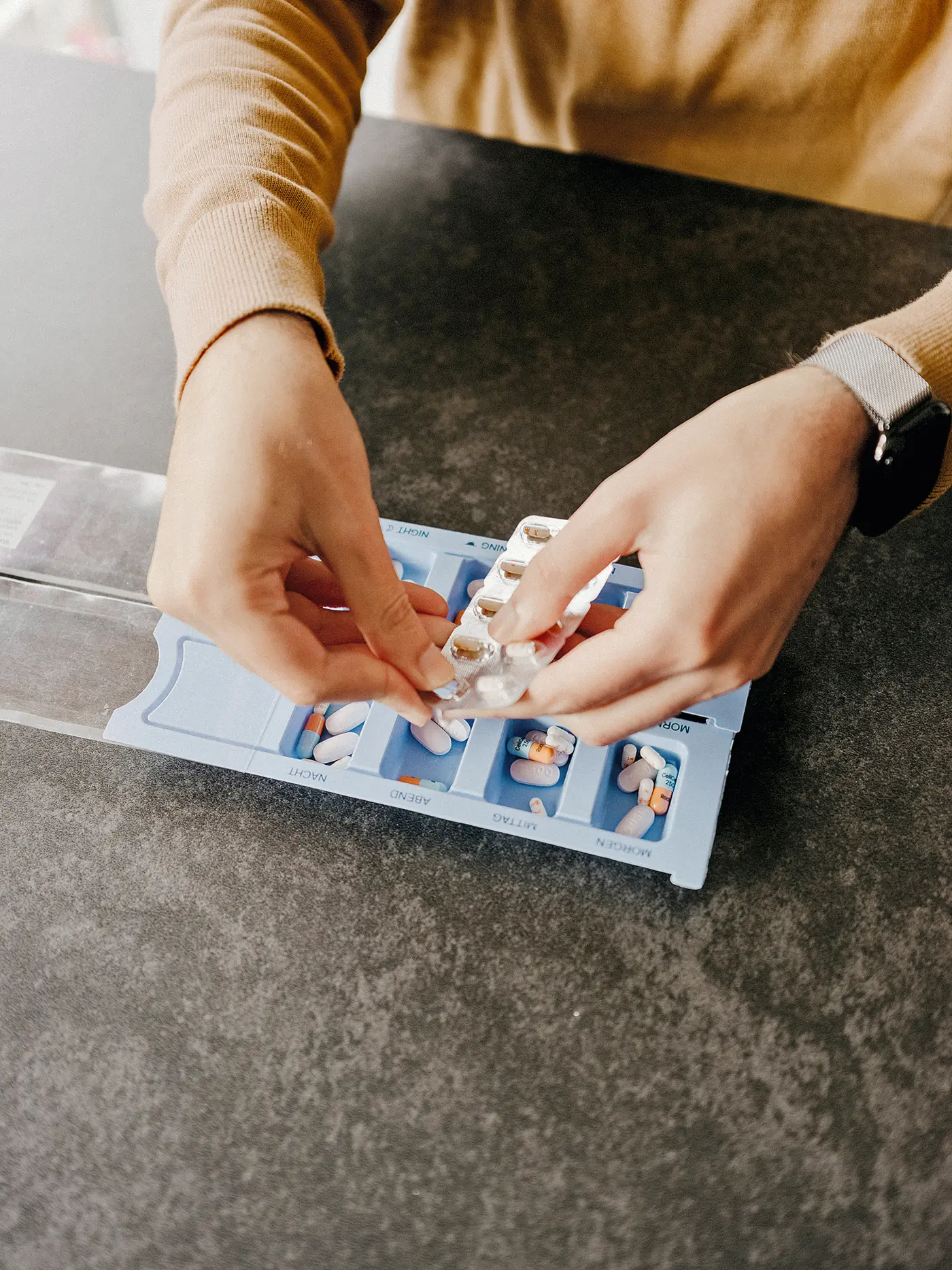 Hände sortieren Tabletten aus einer Blisterpackung in eine beschriftete Medikamentenbox mit Tagesfächern.