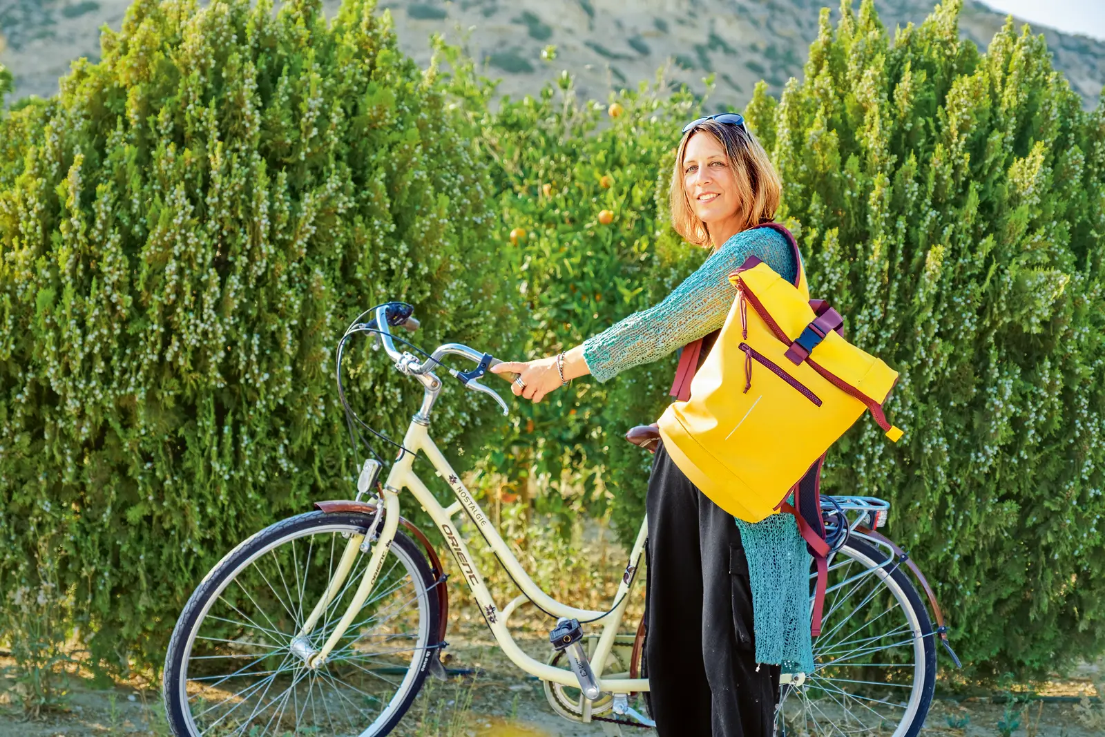 Eine Frau steht neben einem Fahrrad im Grünen, trägt eine Strickjacke und einen gelben Rucksack. Im Hintergrund sind Büsche und Hügel zu sehen, die Sonne scheint.