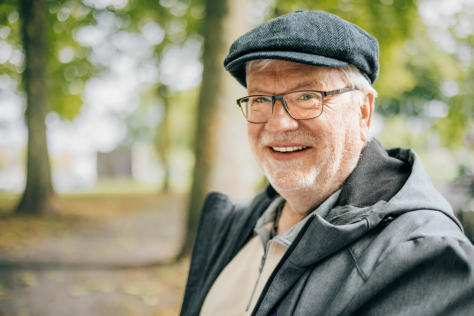Ein älterer Mann mit Brille und grauem Bart lächelt freundlich in die Kamera. Er trägt einen grauen Hut und eine graue Jacke und steht in einem Park mit Bäumen im Hintergrund.