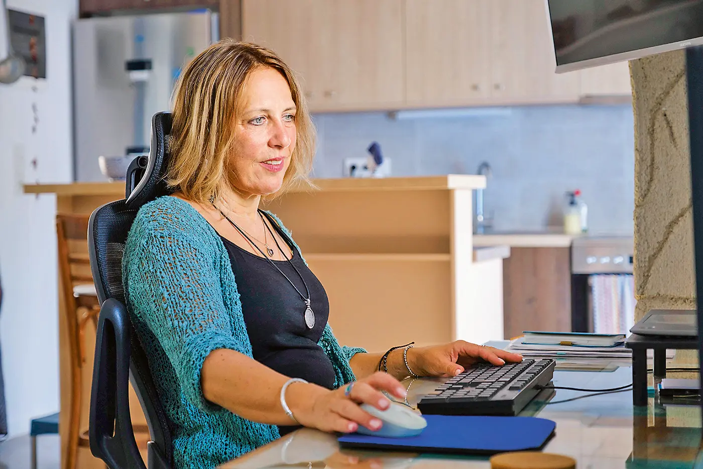 Eine Frau sitzt an einem Schreibtisch in einer modernen Küche und arbeitet am Computer. Sie trägt ein schwarzes Top und eine türkisfarbene Strickjacke, wirkt konzentriert und entspannt.