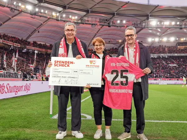 Drei Personen stehen im Stadion auf dem Feld und halten einen Spendencheck in der Hand.