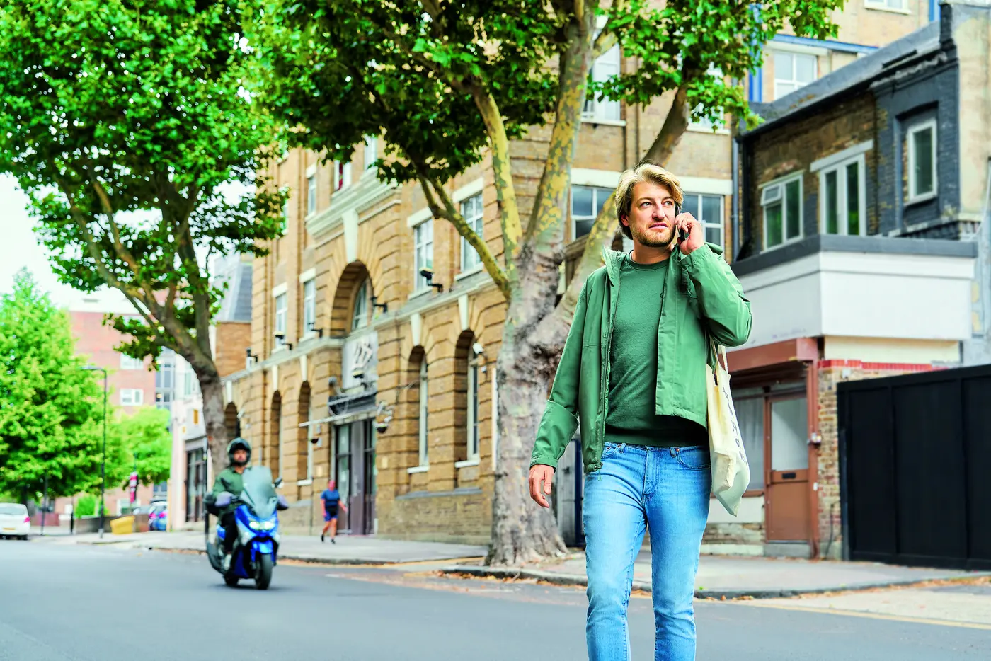Ein Mann steht draußen vor einem Gebäude und spricht am Handy. Er trägt einen grünen Parka, ein grünes T-Shirt und Jeans. Im Hintergrund sind ein Baum und eine Häuserwand zu sehen.