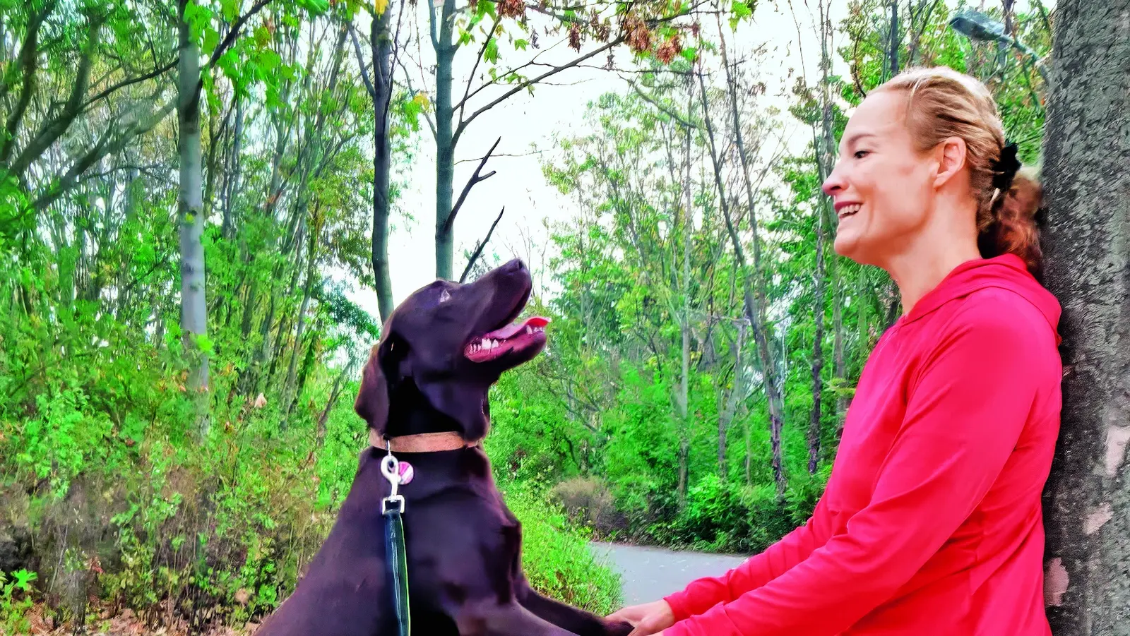 Eine Dame spielt mit ihrem Hund in einem Waldgebiet und lehnt sich dabei mit ihrem Rücken an einen Baum.