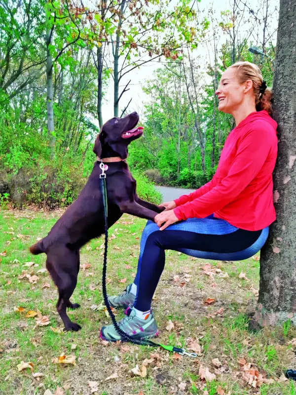 Eine Dame spielt mit ihrem Hund in einem Waldgebiet und lehnt sich dabei mit ihrem Rücken an einen Baum.