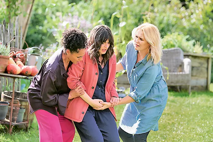 Drei Frauen stehen im Freien, wobei zwei von ihnen eine dritte Frau unterstützen, die anscheinend gestützt wird. Sie wirken fürsorglich und helfen der anderen, die in der Mitte steht, indem sie ihre Arme halten. Die Frauen tragen bunte, lässige Kleidung und stehen in einem grünen Garten