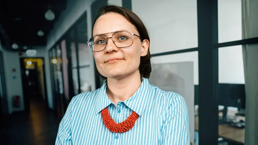 Eine Frau mit Brille und roter Kette steht mit verschränkten Armen in einem Büroflur und blickt selbstbewusst in die Kamera.