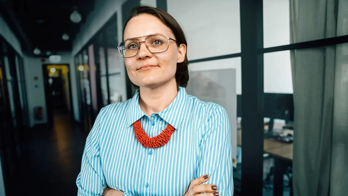 Eine Frau mit Brille und roter Kette steht mit verschränkten Armen in einem Büroflur und blickt selbstbewusst in die Kamera.