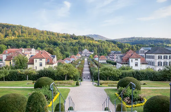 Blick auf die Bäderarchitektur in Bad Brückenau. Eine breite, gepflasterte Allee führt durch den Park, gesäumt von Bäumen und gepflegten Beeten. Im Hintergrund sind mehrere historische Gebäude mit roten Dächern zu sehen, die von einer bewaldeten Hügelkette umgeben sind. Der Himmel ist klar und blau.