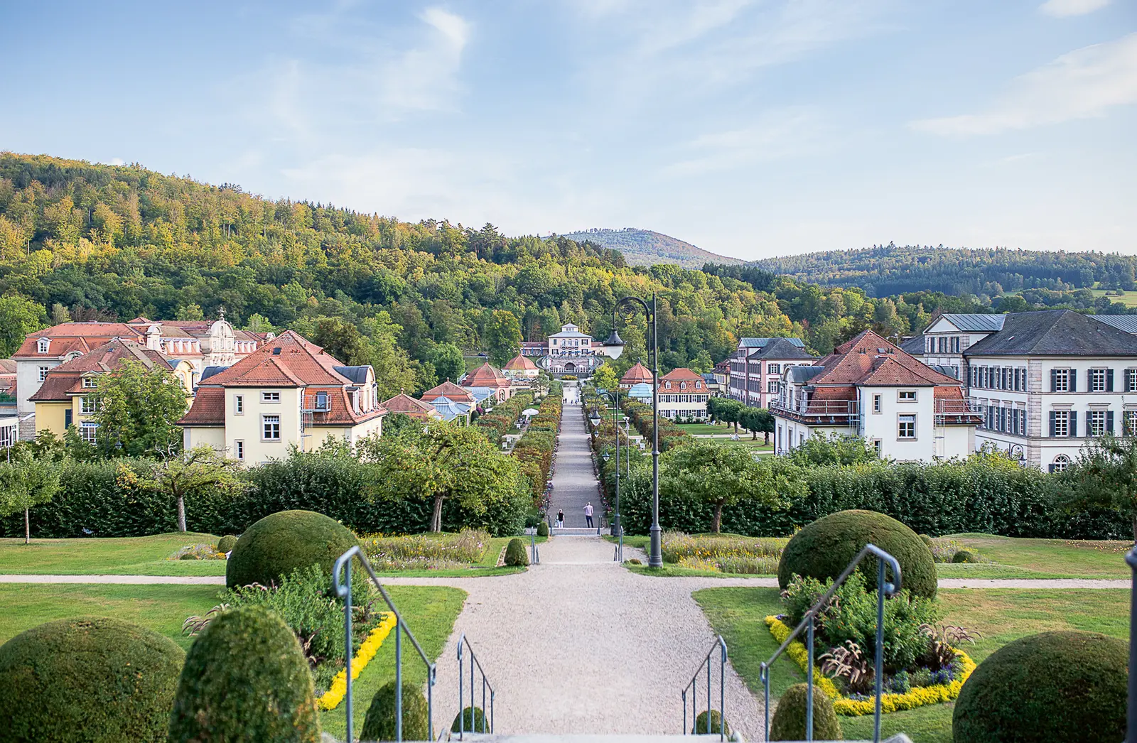 Blick auf die Bäderarchitektur in Bad Brückenau. Eine breite, gepflasterte Allee führt durch den Park, gesäumt von Bäumen und gepflegten Beeten. Im Hintergrund sind mehrere historische Gebäude mit roten Dächern zu sehen, die von einer bewaldeten Hügelkette umgeben sind. Der Himmel ist klar und blau.