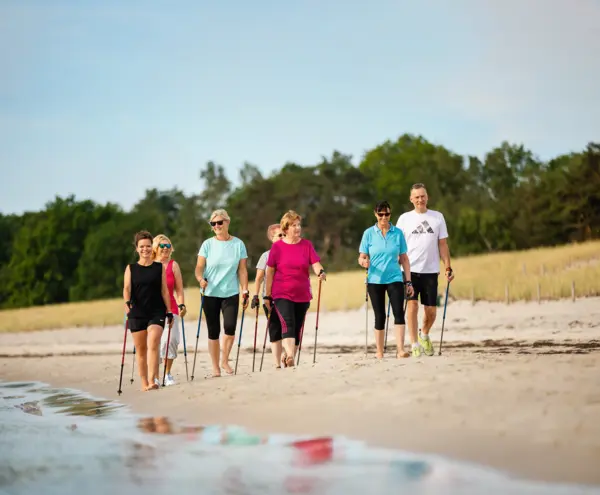 Eine Gruppe von Männern und Frauen geht gemeinsam mit Nordic-Walking-Stöcken am Strand entlang, im Hintergrund Dünen und Bäume.