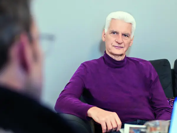 Ein älterer Mann sitzt auf einem Sofa und blickt direkt in die Kamera.