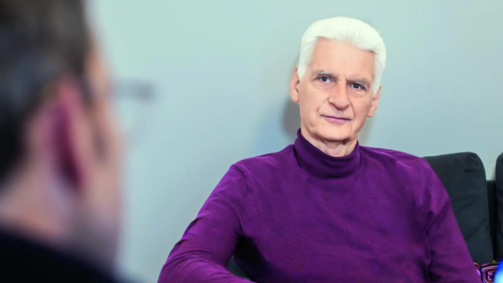 Ein älterer Mann sitzt auf einem Sofa und blickt direkt in die Kamera.