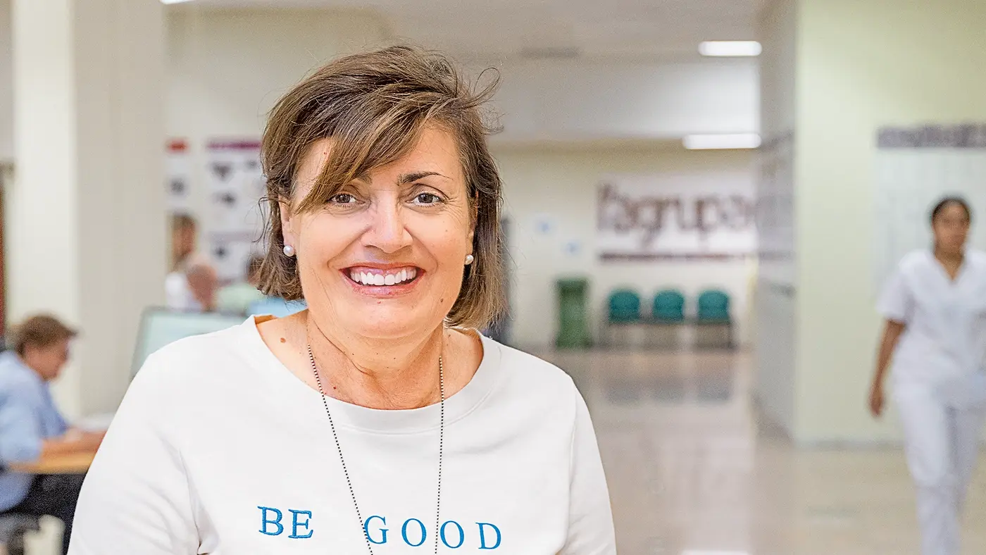 Eine lächelnde Frau mit kurzem braunem Haar trägt ein weißes Shirt. Im Hintergrund sind weitere Personen in einem hellen Raum zu sehen, an der Wand hängen Fotos und Poster.