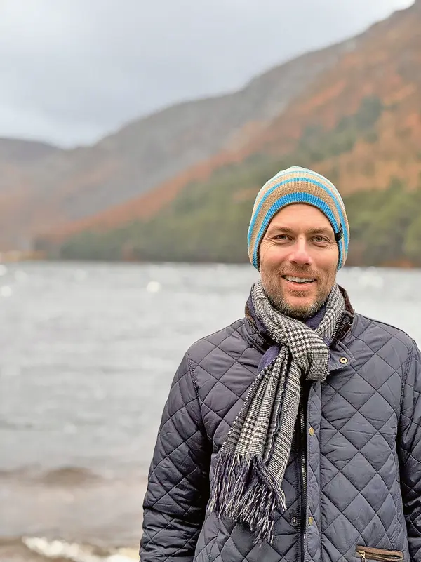 Ein Mann steht im Freien vor einem See und trägt einen bunten Mütze sowie einen grauen Schal. Er hat ein breites Lächeln auf dem Gesicht und trägt eine dunkelblaue Jacke. Im Hintergrund sind Berge und Wälder zu sehen.