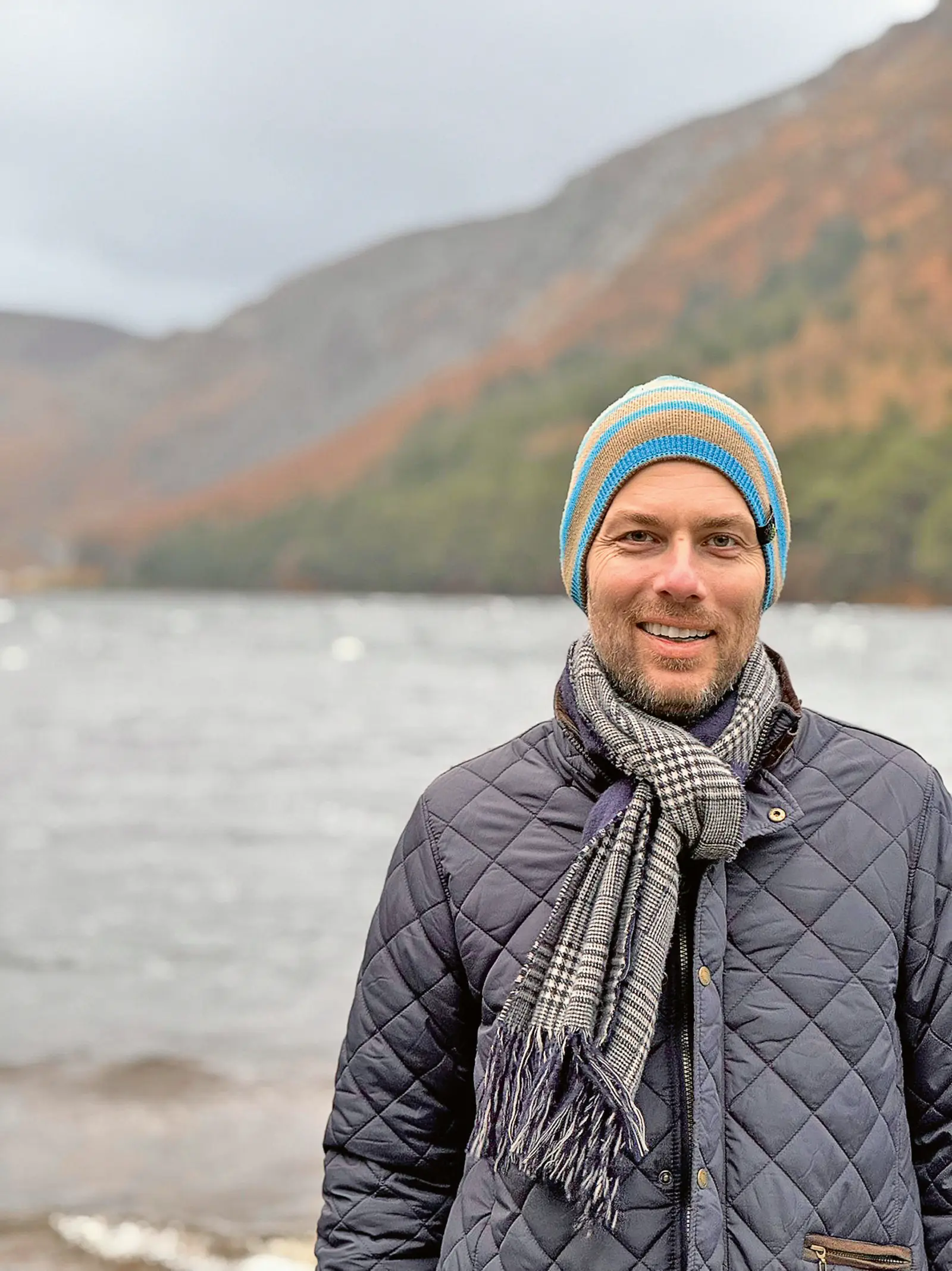Ein Mann steht im Freien vor einem See und trägt einen bunten Mütze sowie einen grauen Schal. Er hat ein breites Lächeln auf dem Gesicht und trägt eine dunkelblaue Jacke. Im Hintergrund sind Berge und Wälder zu sehen.