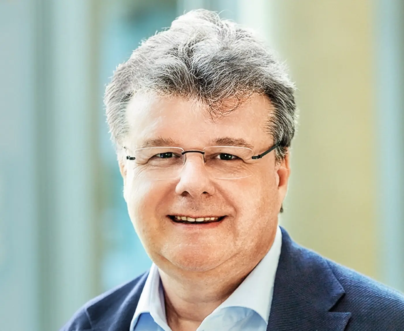 Ein Mann mit grauen Haaren und Brille lächelt in die Kamera, trägt Hemd und Sakko vor hellem Hintergrund.