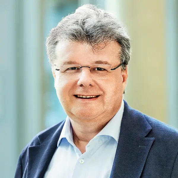 Ein Mann mit grauen Haaren und Brille lächelt in die Kamera, trägt Hemd und Sakko vor hellem Hintergrund.