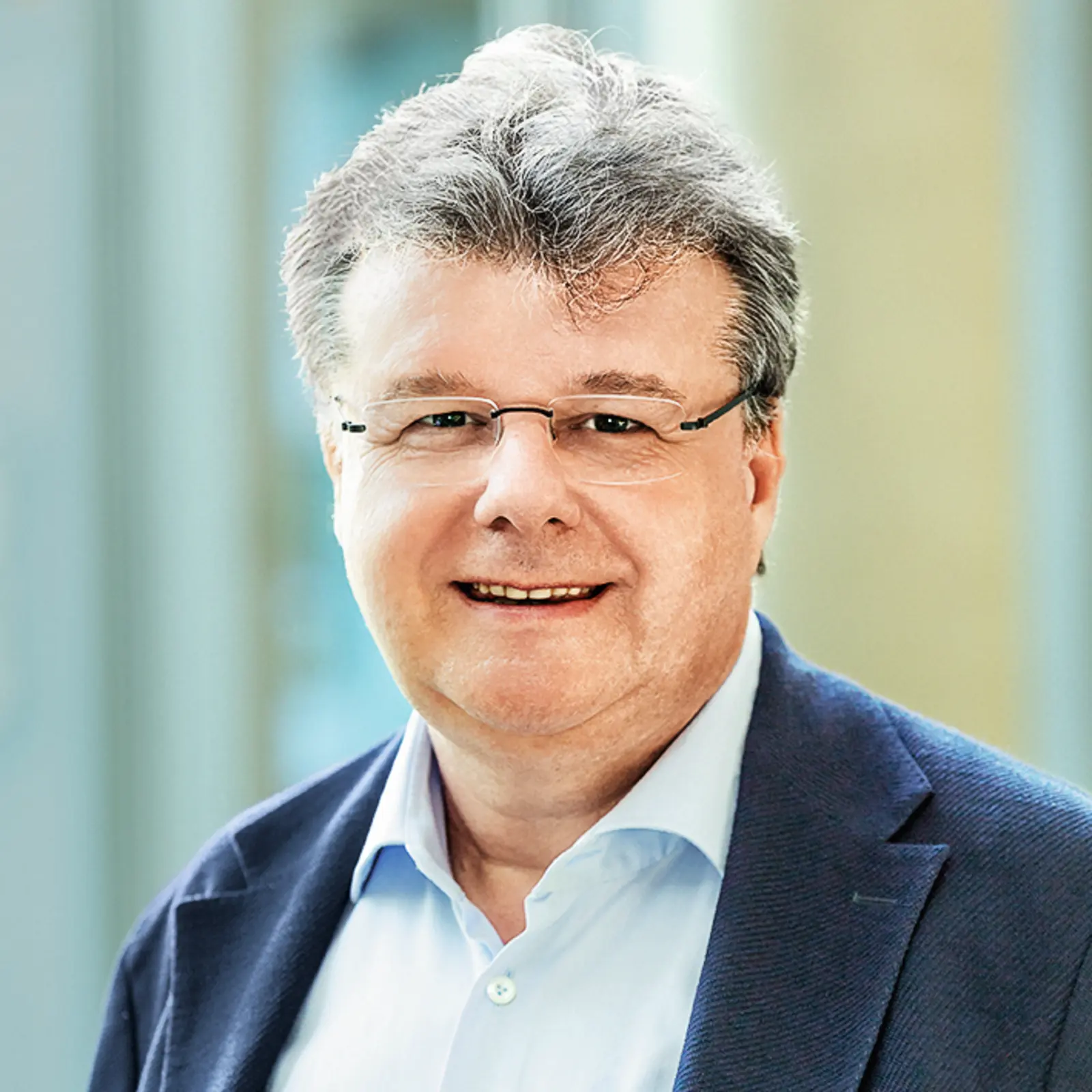 Ein Mann mit grauen Haaren und Brille lächelt in die Kamera, trägt Hemd und Sakko vor hellem Hintergrund.