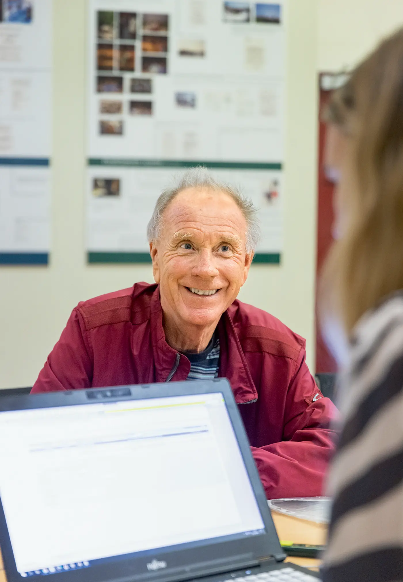 Ein älterer Mann mit grauen Haaren und roter Jacke sitzt lächelnd an einem Tisch. Ihm gegenüber sitzt eine Frau, vor der ein Laptop steht. Im Hintergrund hängen Pinnwände mit Fotos und Unterlagen.