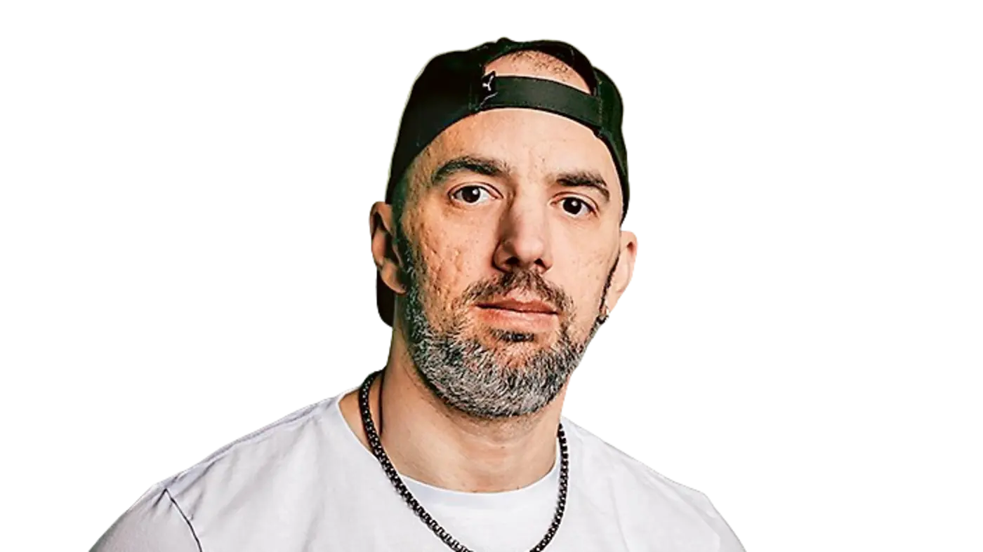 ChatGPT: Porträt eines Mannes mit kurzem Bart und grauen Haaren, der eine schwarze Basecap rückwärts trägt. Er schaut ernst in die Kamera, trägt ein weißes T-Shirt und eine silberfarbene Gliederkette.