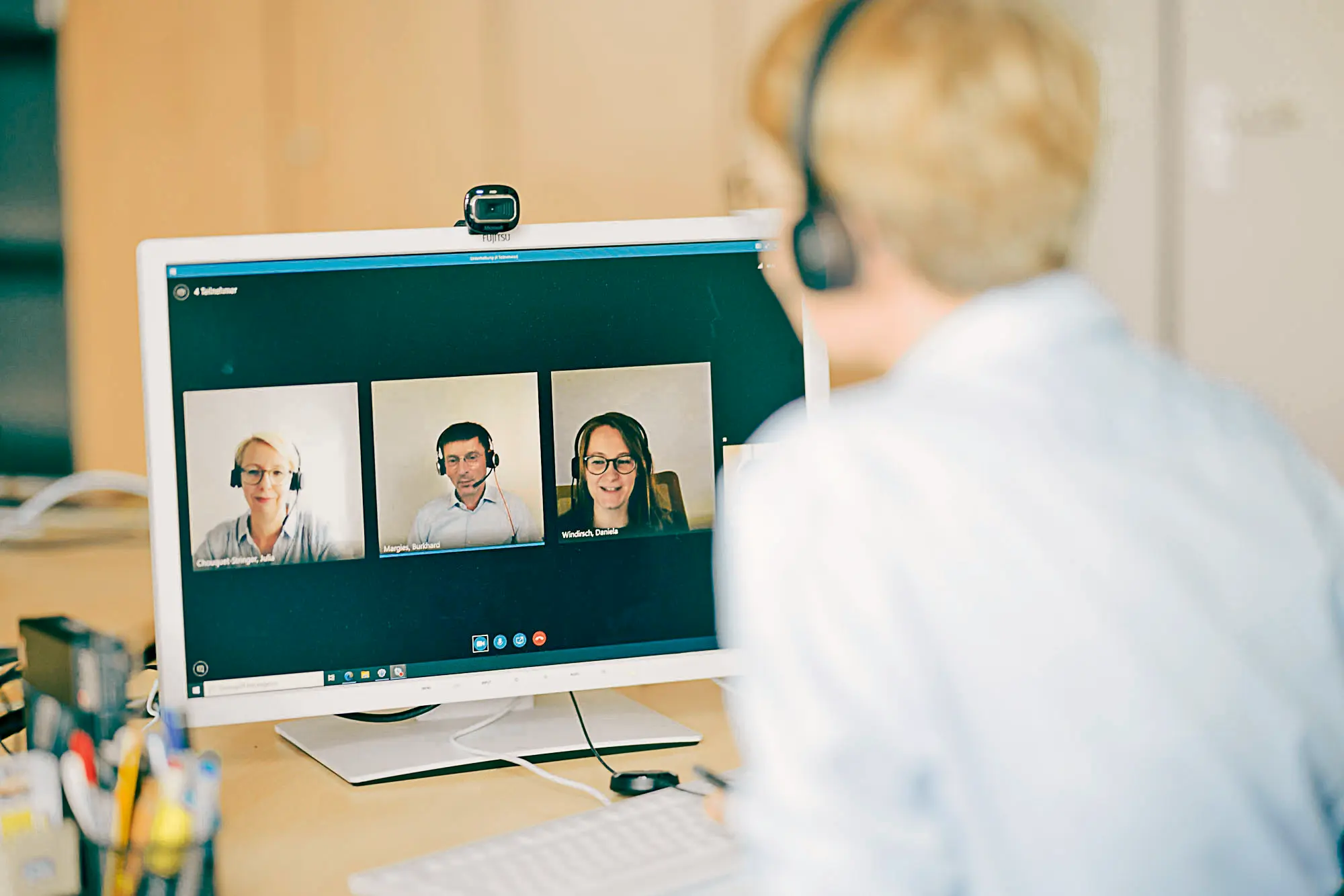 ChatGPT:  Eine Person mit Headset sitzt am Schreibtisch und nimmt an einer Videokonferenz teil. Auf dem Monitor sind drei Gesprächspartnerinnen und Gesprächspartner in separaten Fenstern zu sehen.