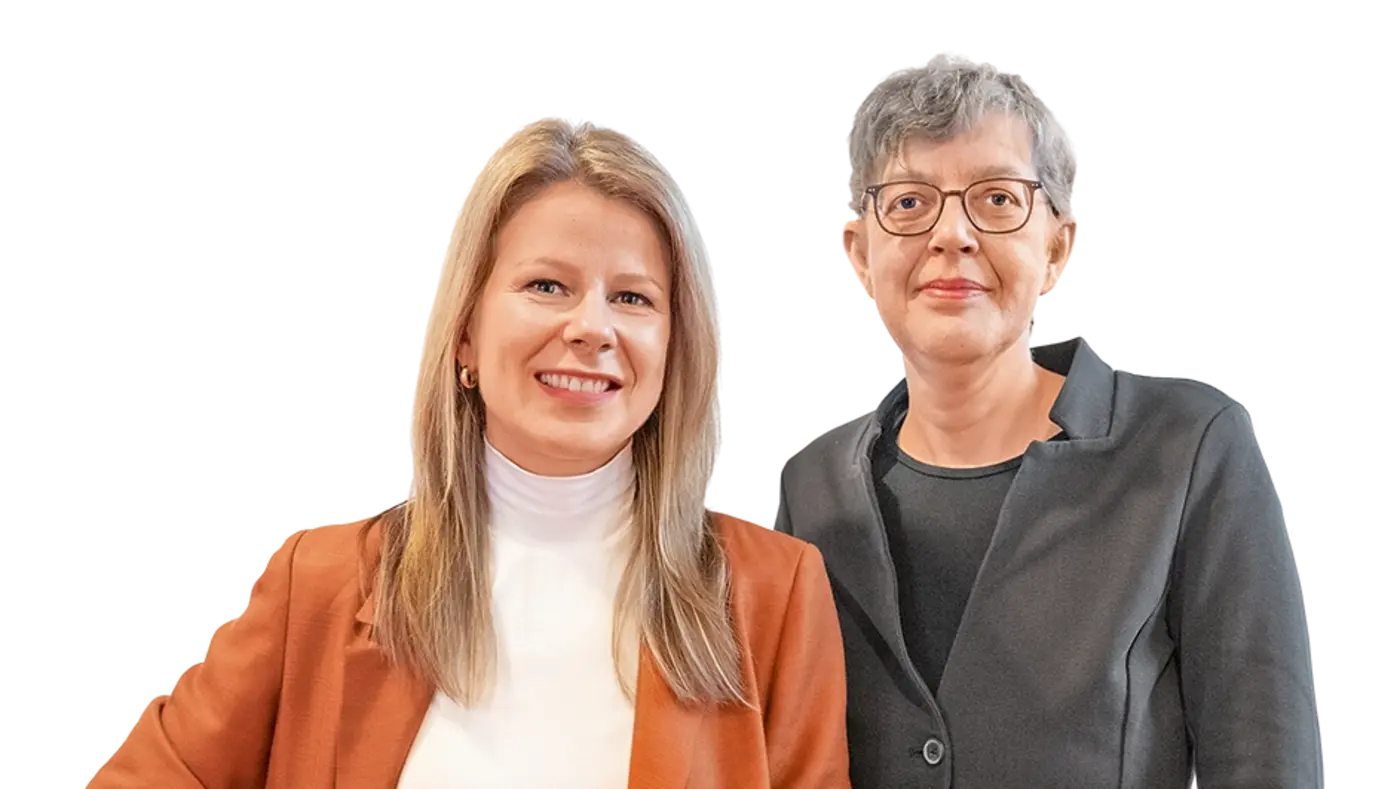 Das Bild zeigt zwei Frauen, die nebeneinander stehen. Die Frau auf der linken Seite trägt einen orangenen Anzug und ein weißes Oberteil, während die Frau auf der rechten Seite ein schwarzes Jackett und eine schwarze Bluse trägt. Beide blicken in die Kamera und stehen in einem professionellen, aber entspannten Pose.