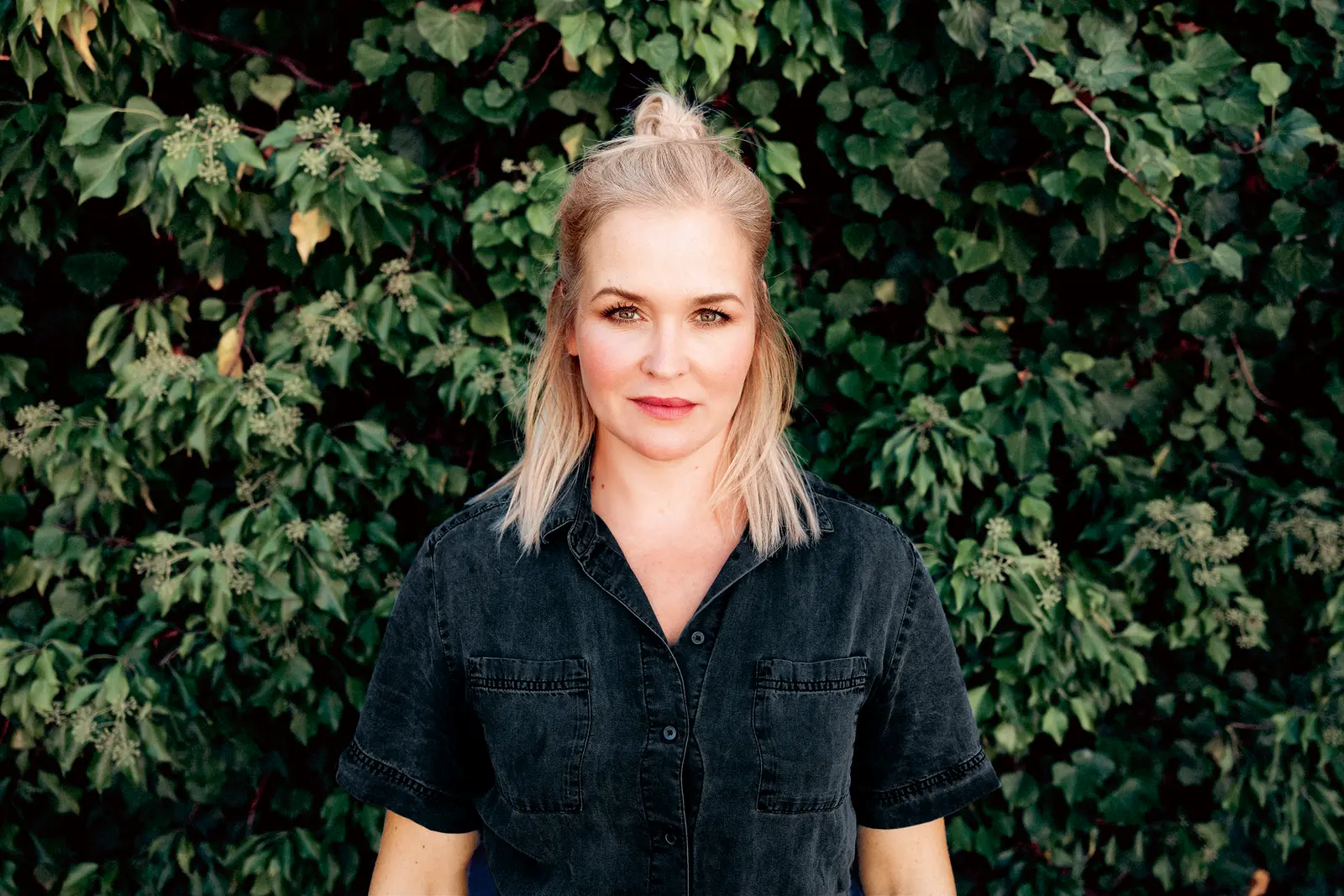 Porträt einer Frau mit blonden, schulterlangen Haaren, die in einem hohen Dutt gebunden sind. Sie trägt ein schwarzes Hemd und steht vor einem grünen, pflanzenbewachsenen Hintergrund. Sie schaut direkt in die Kamera und hat einen ernsten, aber selbstbewussten Ausdruck.
