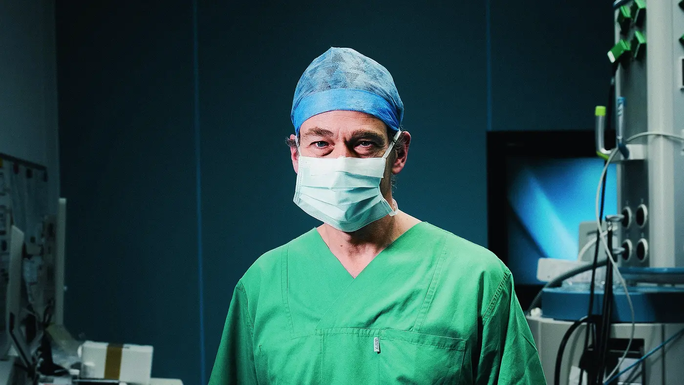 Ein Chirurg in grüner OP-Kleidung und Mundschutz steht im Operationssaal, umgeben von medizinischen Geräten.