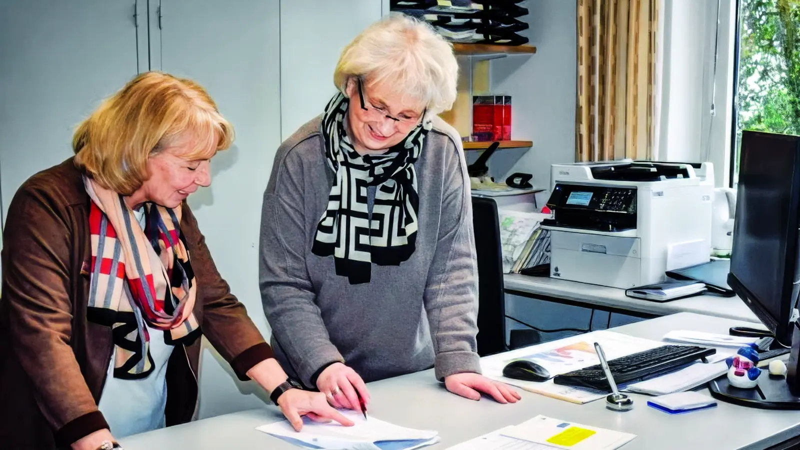 Zwei ältere Frauen sind in einem Büro an einem Schreibtisch und betrachten gemeinsam Unterlagen.