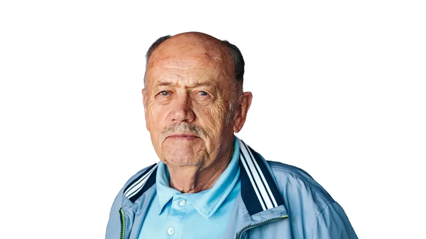 Freigestelltes Porträt eines älteren Mannes im hellblauen Poloshirt und blauer Jacke vor weißem Hintergrund.