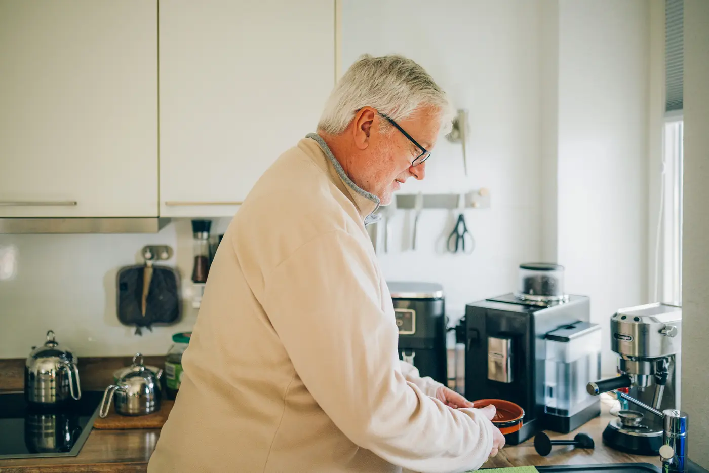 Ein älterer Mann steht in einer modernen Küche und bereitet etwas vor. Er hält eine kleine Schüssel in der Hand und schaut konzentriert auf das, was er tut. Hinter ihm sind verschiedene Küchengeräte sichtbar.