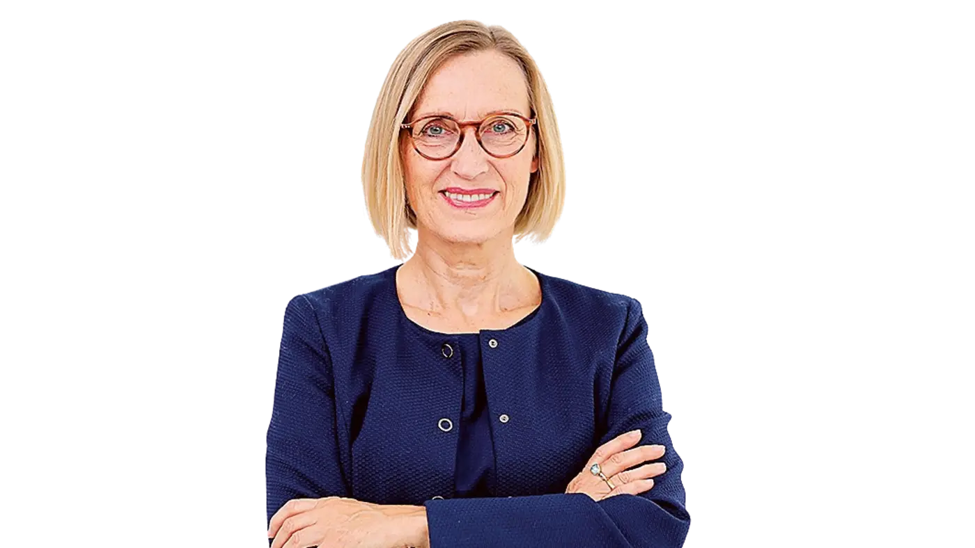 Porträt einer Frau mit kinnlangem blonden Haar und Brille, die in die Kamera lächelt. Sie trägt ein dunkelblaues Jackett und hat die Arme locker vor der Brust verschränkt.