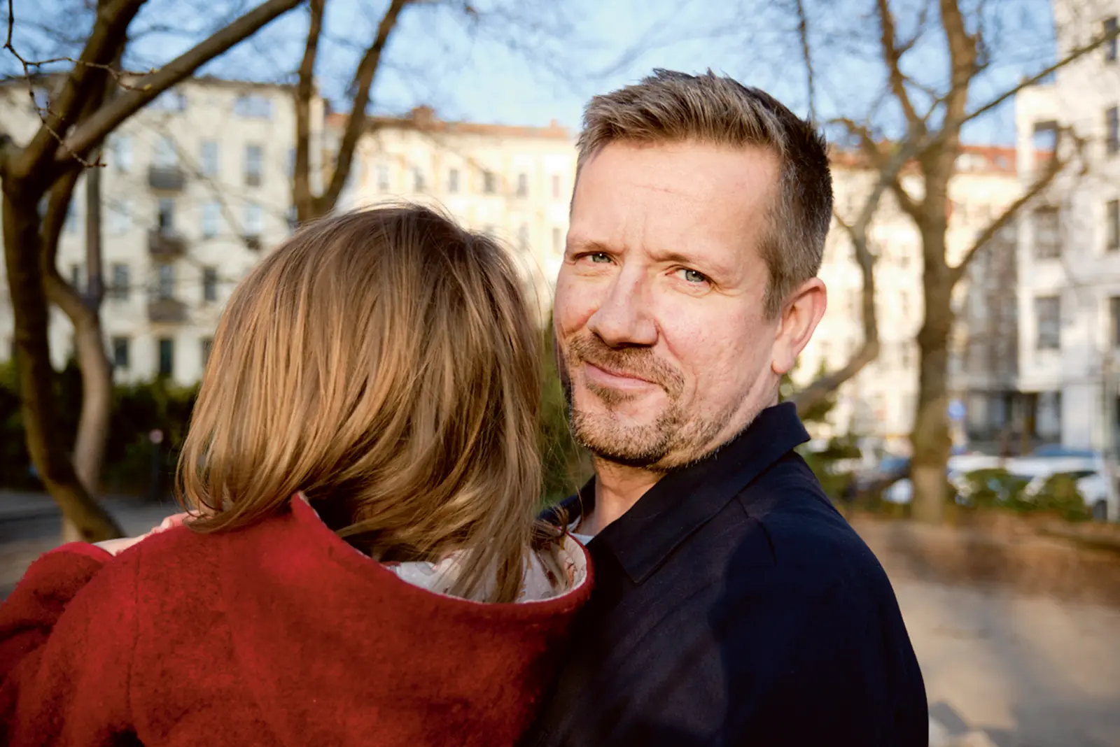 Ein Mann mit kurzem Haar und Bart steht im Freien und hält ein Kind im Arm, das den Kopf an seine Schulter lehnt und einen roten Mantel trägt. Der Mann blickt mit leichtem Lächeln in die Kamera. Im Hintergrund sind unscharf Wohnhäuser und kahle Bäume zu sehen.