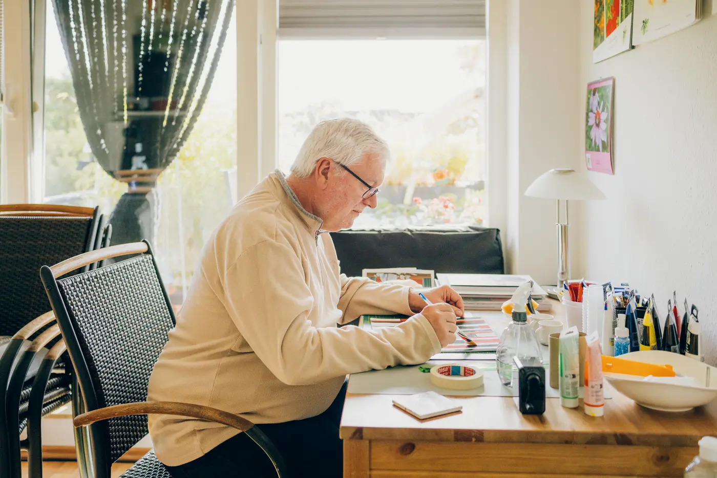 Ein älterer Mann sitzt an einem Holztisch in einem gut beleuchteten Raum und arbeitet mit Buntstiften und Papier. Auf dem Tisch sind verschiedene Kunstmaterialien wie Stifte, Kleber und Notizblöcke zu sehen.