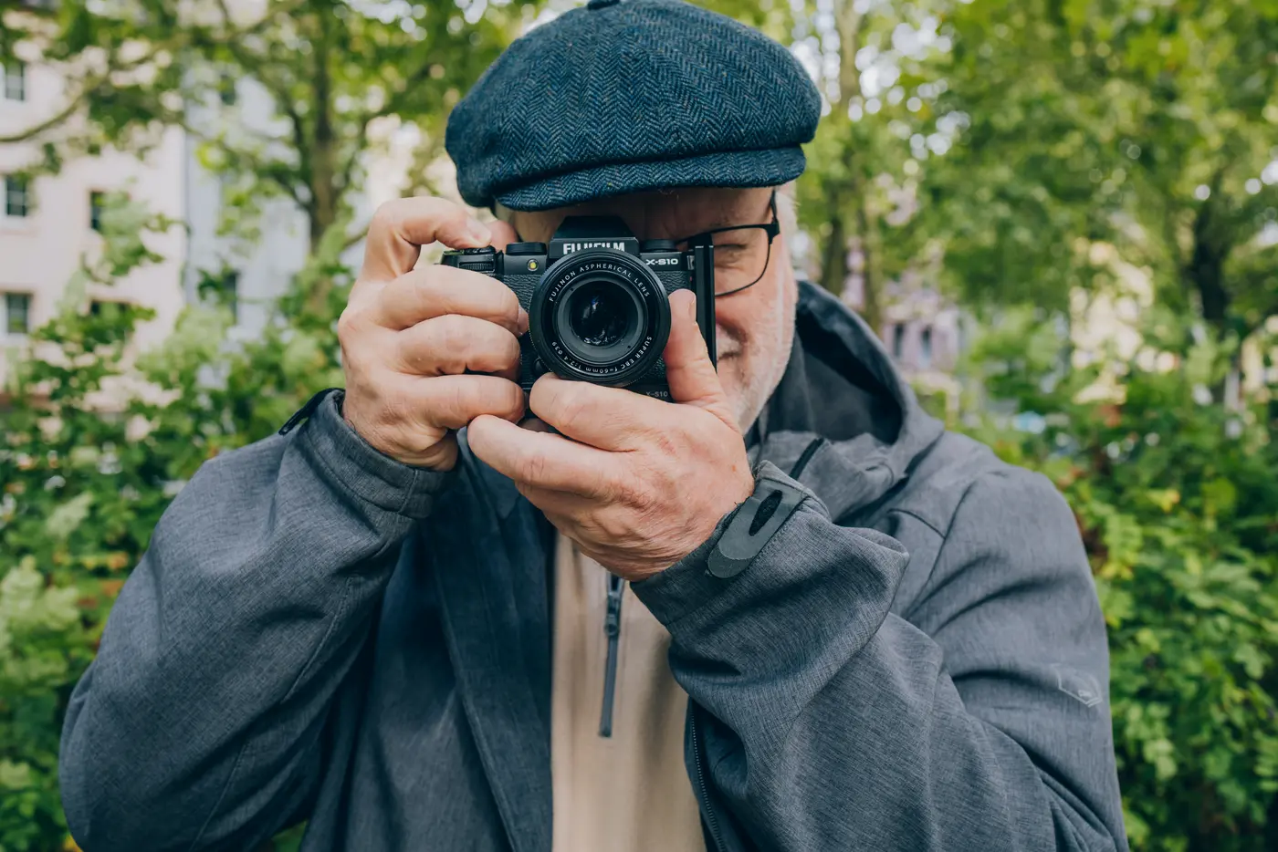 Ein älterer Mann mit Brille und Mütze steht draußen und hält eine Kamera in der Hand. Er schaut durch das Objektiv, während er das Bild aufnimmt.