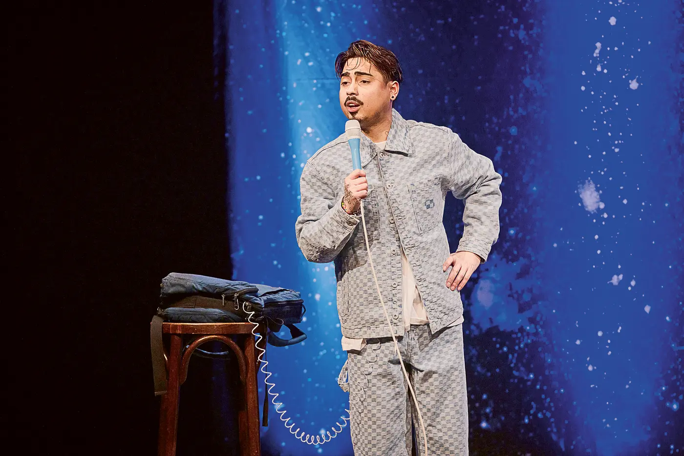 Ein junger Comedian steht auf einer Bühne mit einem Mikrofon in der Hand. Er trägt ein gemustertes, helles Set aus Jacke und Hose. Im Hintergrund ist ein blauer Vorhang mit Sternenmustern zu sehen, der an eine Galaxie erinnert. Neben ihm steht ein Hocker mit einem zusammengefalteten Fallschirm-Rucksack darauf, von dem ein Kabel zum Mikrofon führt. Der Künstler spricht mit ausdrucksvoller Mimik ins Mikrofon, die rechte Hand in die Hüfte gestemmt.