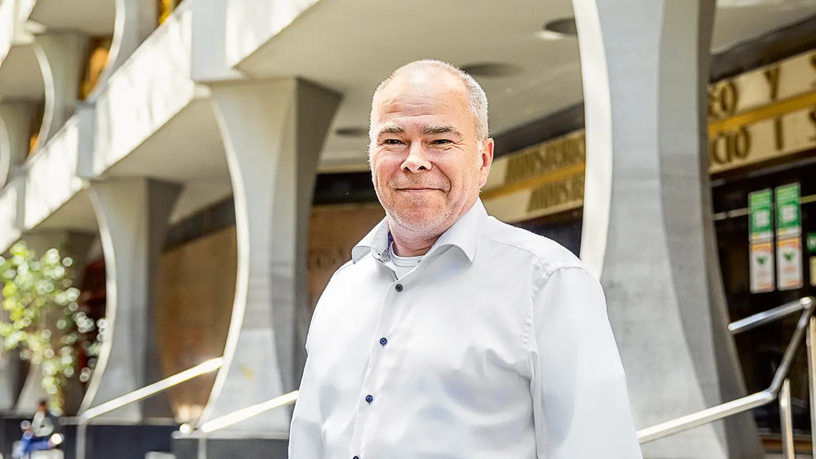 Ein Mann mit kurzem grauem Haar und weißem Hemd steht lächelnd vor einem modernen Gebäude mit geschwungenen Säulen und wehenden Flaggen. Im Hintergrund ist eine sonnige Straßenkulisse zu sehen.